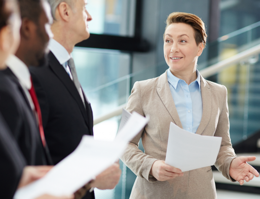woman holding paper, speaking to group of businessmen